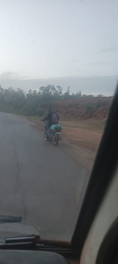 A blurry, low-resolution photo taken from inside a vehicle shows a person riding a motorcycle on a road. The motorcycle rider is wearing dark clothing and carrying a green bag. The road runs alongside a dirt embankment covered with sparse vegetation. Trees are visible in the background. The sky is overcast. A portion of the vehicle's interior is visible in the foreground. The location is presumed to be Mangu Halle, Nigeria.