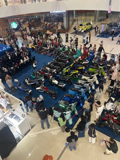 The image depicts a bustling indoor event, likely a mall or exhibition center, filled with people and a display of motorcycles and scooters. The event appears to be taking place in Dumai City, Indonesia, suggested by a visible Starbucks sign, a popular international chain with a presence in the region.
The scene is a high-angle shot looking down on a large, open space with a blue carpeted floor. In the background, there are storefronts, including a prominent Starbucks with its iconic green logo. Beyond the Starbucks, a light green car is parked on a display platform, and next to it, a dark SUV. Various seating arrangements, including black chairs and tables, are visible throughout the area.
The foreground and middle ground are dominated by a large collection of motorbikes and scooters, parked in rows on the blue carpet. The vehicles are varied in color and style, with many modern scooters, some vintage-looking models, and several sportier motorcycles. A number of people are gathered around the vehicles, some examining them closely, others chatting amongst themselves. Many attendees are dressed in casual attire, with a notable presence of individuals wearing hijabs, consistent with the cultural context of Indonesia.
The crowd is diverse, with men, women, and children present. Some individuals appear to be engaged in conversations, while others are taking photographs or videos of the display. The atmosphere seems lively and engaging, with people actively participating in the event.
Specific actions observed include individuals standing and looking at the vehicles, people walking through the aisles between the motorcycles, and some individuals gathered in small groups. One person in the foreground is seen taking a picture with their phone, while another is seated on the floor with a plastic bag. A few individuals are interacting with what appear to be information booths or merchandise stands. The overall impression is of a public gathering centered around a motorcycle exhibition.