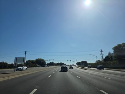 This media file captures a wide-angle view of a multi-lane road in Elk Grove, USA, under a clear, bright blue sky. The sun is high in the sky, suggesting it is midday. The weather appears to be sunny and warm.
The primary subjects are the road and the vehicles on it, indicating a scene of everyday travel. Several cars are visible, moving in the same direction as the camera. In the foreground, the asphalt road is marked with white lane lines. To the left, there is a sidewalk and a median with some vegetation and a large, blank sign. Power lines and utility poles run along the side of the road, and a dense line of trees and bushes borders the landscape. In the distance, more trees and some buildings are visible. High in the sky, a faint white contrail from an airplane stretches across the blue expanse.
There is no discernible activity or event taking place other than vehicles traveling on the road. The scene conveys a sense of ordinary daytime transit. No people are clearly visible within the vehicles.
Notable details include the bright sunlight, casting a slight glare in the upper right portion of the image, and the clear blue sky with the airplane contrail. The location is identified as Elk Grove, United States of America. There is no emotionally charged content or specific atmospheric mood conveyed.
Visible text includes a road sign on the right side of the road, partially obscured by trees, with the number "99" clearly visible, likely indicating a highway number. Another sign on the left side is blank and appears to be a directional or informational sign.
