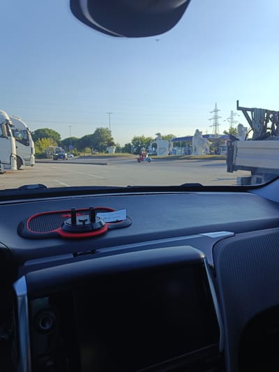 The image shows a view from inside a car, looking out through the windshield. The dashboard is in the foreground, with a black and red phone mount and a small piece of paper on it. The paper has some handwritten text in what appears to be Italian. Beyond the car, the scene is a parking lot or rest stop.
In the middle ground, there is a gas station with large, abstract white sculptures in front of it. There are several vehicles, including a large white truck, a smaller black car, and a red scooter. Trees and utility poles are visible in the background.
The sky is clear and blue, suggesting it is daytime, likely morning or late afternoon due to the low angle of the sun casting long shadows. The weather appears to be fair. The overall setting is a roadside service area. No people are clearly identifiable in the image.
The visible text on the paper appears to be a date and time, possibly a receipt or note. It reads "21/05/24 12:01".