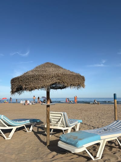 The image depicts a sandy beach in Benicàssim, Spain, under a clear blue sky with faint cirrus clouds. In the foreground, two white chaise lounges with blue and white striped cushions are positioned on the sand, with one prominently featuring a light blue cushion on its base. A large thatched umbrella, supported by a wooden pole, partially covers the chaise lounges. A circular wooden table is attached to the umbrella's pole.
In the midground, several people are visible on the beach. Some are standing and interacting, while others are seated on beach chairs or lying on towels. Several colorful beach umbrellas are scattered across the sand, and the gentle waves of the Mediterranean Sea are visible in the background. The overall scene is one of a tranquil beach environment, likely during daylight hours.