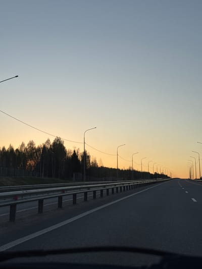 The image shows a highway in Ivanovka, Russia at dusk or dawn. The road is asphalt and has white lane markings. A metal guardrail runs along the side of the road, separating it from a line of trees. Streetlights are evenly spaced along the road, extending into the distance. The sky transitions from blue to orange as the light fades. The image appears to be taken from a moving vehicle.