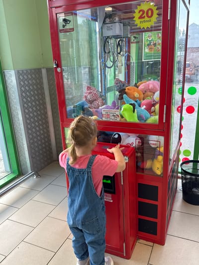 This vertical, outdoor, medium shot captures a young girl playing with a claw machine filled with plush toys. The girl, wearing a pink shirt and denim overalls, is reaching towards the machine's controls. The claw machine is bright red and has a glass front displaying various colorful stuffed animals. A sign on the machine indicates that a game costs 20 rubles. There are also signs in Russian, with one warning that video surveillance is in progress. The setting appears to be a public area with tiled flooring and a metal railing. The lighting suggests it's daytime, with natural light illuminating the scene. The girl's focused expression suggests she is engrossed in the game. The overall atmosphere is playful and engaging.
