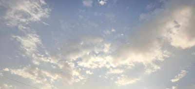 The image captures a sky filled with clouds over Margilan City, Uzbekistan. The sky is a pale blue, transitioning to a lighter hue near the horizon, indicating either early morning or late afternoon. Scattered white and gray clouds of varying sizes and densities drift across the frame. Some clouds are thin and wispy, while others are more substantial and opaque, catching the light from the sun which is not directly visible but its presence is suggested by the illumination of the cloud edges. A single, thin black wire stretches diagonally across the lower left portion of the image, possibly a power line or telephone wire, indicating the presence of human infrastructure. The overall scene is serene and atmospheric, depicting a typical sky over a city.