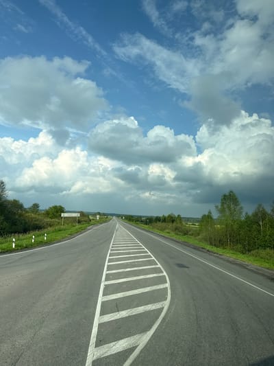 The image depicts an asphalt road curving into the distance under a partly cloudy blue sky. The road is marked with white lines, including a prominent series of diagonal lines on the right side, suggesting a lane separation or merging area. White roadside markers with black stripes are visible along the left edge of the road. The landscape flanking the road consists of green grass, bushes, and scattered trees. In the far distance, the road continues, framed by a flat, tree-lined horizon under a dramatic sky filled with large, fluffy white and gray clouds. The scene appears to be a rural or semi-rural setting.