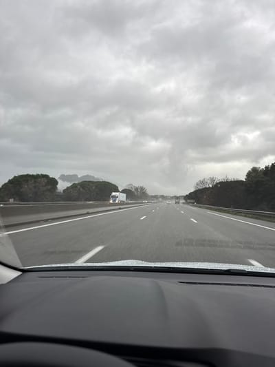 A highway stretches into the distance under a heavy, overcast sky. The road surface is wet, reflecting the dim light. A few vehicles are visible ahead, including a semi-truck on the left. Trees line the sides of the highway, their silhouettes stark against the gray backdrop. The shot is taken from inside a car, with part of the dashboard visible on the right, raindrops clinging to the window.