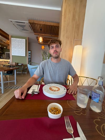A young man with curly brown hair and a beard is seated at a wooden table in a restaurant. He is wearing a grey t-shirt. In front of him is a white bowl containing a tomato-based soup topped with cheese or breadcrumbs. To the right of the soup, there is a glass of water and a partially full bottle of water. A small white bowl filled with nuts is also on the table. The background shows a ceiling with an air conditioning unit and wooden beams, as well as lamps and what appears to be a bar area with the sign "GASTROBAR" visible. The restaurant is likely located in Mojácar, Spain, given the context. The man's left hand rests on the table near the soup.