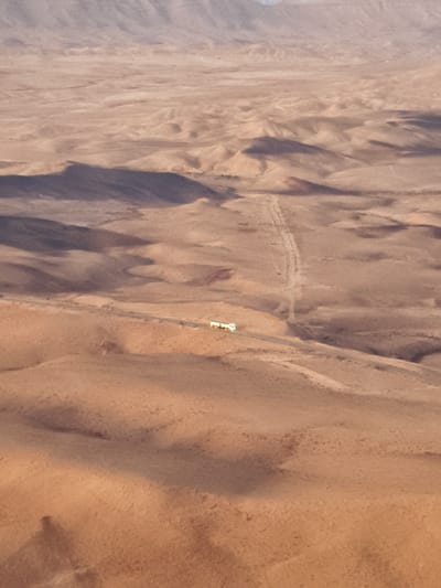 An aerial view shows a vast, undulating desert landscape. The terrain is characterized by sand dunes and barren, rocky hills, with subtle variations in color ranging from light tan to darker brown, creating a sense of depth and texture. Shadows are cast across the landscape, indicating a low sun angle. A paved road cuts across the middle ground of the image, winding through the desert. On this road, a white bus with yellow accents is visible, appearing to travel from left to right. In the distance, the hazy outlines of mountains are visible against a pale sky. The scene is devoid of vegetation. This image depicts the Negev Desert, likely near Yeruham, Israel.