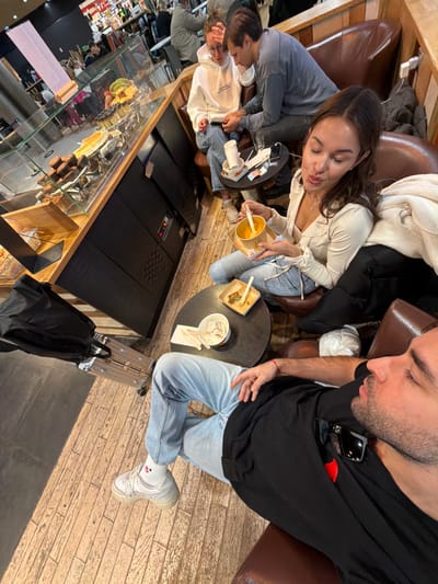 The scene depicts four individuals seated in a cafe or food court within Colombier-Saugnieu, France, likely at Lyon-Saint Exupéry Airport.
In the foreground, a male with dark hair and a beard, wearing a black t-shirt with a red heart emblem and light blue jeans, is seated in a brown upholstered armchair. He looks towards the left with his eyes partially closed, his right hand resting on his thigh. White socks with a red graphic and white sneakers are visible. Sunglasses are tucked into his shirt collar. To his immediate left, a black backpack rests on a silver-colored rolling suitcase with an extended handle.
In the mid-ground, a female with long dark hair, wearing a light-colored long-sleeved top and light blue jeans, is seated in a brown upholstered chair opposite a dark circular table. She holds a cardboard bowl containing an orange-colored food item with a wooden spoon, bringing the spoon towards her mouth. Her lips are slightly puckered. A white jacket is draped over the back of her chair. On the table, an empty cardboard bowl with a wooden utensil and a crumpled napkin are present.
In the background, two individuals occupy brown upholstered seating. A female with light brown hair and glasses, wearing a white hooded sweatshirt and blue jeans, is seated, looking down at a mobile phone she holds. Beside her, a male with short dark hair, wearing a grey long-sleeved shirt and dark pants, leans towards her, also looking down, potentially at the same device. A white paper cup, a mobile phone, and some banknotes are on the dark circular table in front of them.
To the left of the image, a food service counter features a glass display case showcasing various baked goods, including rows of muffins, a large tart, and fresh fruit such as bananas. The counter base is dark-colored. The floor throughout the seating area is composed of light brown wooden planks, transitioning to a darker, smooth surface near the luggage. Wooden paneling lines the wall behind the seated individuals, and the area is illuminated by artificial lighting.