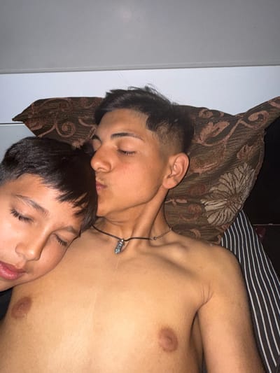 An overhead shot captures two young individuals, likely male, with dark hair and medium skin tone, resting closely together in what appears to be an indoor bedroom setting. The image is rotated, presenting the subjects vertically.
The person in the upper half of the frame is lying on a brown pillow with a floral and swirling pattern, their eyes closed as if sleeping. They are shirtless, revealing their upper torso and shoulder, and wear a black cord necklace with a small, clear or blue-toned pendant. Their head is resting on the head of the second person, who occupies the lower half of the frame. This second individual also has their eyes closed and appears to be sleeping or resting, with a faint smile visible on their lips.
Behind the subjects, a striped fabric (black and white) is visible, suggesting a sheet or blanket. A plain, light-colored surface, possibly a wall or headboard, forms the left background. The lighting in the image is dim, indicating either nighttime or a darkened room. There is no visible text. The overall atmosphere conveyed is one of peace, intimacy, and relaxation. No specific location cues are present within the image itself to confirm the provided context of Zlatna Panega, Bulgaria.
