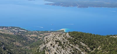 This is a high-angle, wide shot of a coastal landscape in Općina Bol, Croatia. In the foreground, a rocky, tree-covered mountainside slopes down towards a town. The town consists of numerous buildings, many with red tile roofs, nestled among trees along the coastline. The town is situated next to the bright blue Adriatic Sea. Several boats are visible on the water, including a ferry and smaller speedboats leaving white wakes. The sea extends to the horizon, where it meets a hazy shoreline with islands. The overall scene is bathed in bright sunlight, indicative of a clear day.