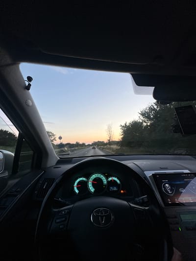 The image is taken from the interior of a car looking forward as it drives down a road at sunset. The dashboard is visible, including the steering wheel with the Toyota logo, the instrument cluster displaying speed and RPM, and a touchscreen infotainment system. Outside the car, the sky is a gradient of orange and blue, indicating the sun is setting. Trees and fields line the road, and other cars are visible in the distance. The scene appears to be in a rural or semi-rural area.