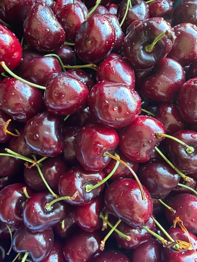 The media file is a close-up, overhead shot of a pile of dark red cherries, glistening with water droplets. The cherries are deep in color, appearing ripe and fresh. They are clustered together, with some stems still attached. The water droplets on the surface of the cherries create a sparkling effect, suggesting they may have been recently washed or are dewy. The lighting appears bright, possibly natural daylight, as indicated by the highlights on the cherries. The scene is a simple and focused one, with the cherries being the sole subject. There is no visible text in the image. The context of "Aespa, Estonia" suggests a possible outdoor setting, perhaps a garden or a farm, where these cherries might have been harvested. The overall impression is one of freshness and abundance of fruit.
