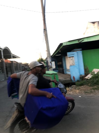 Two men are riding a motorcycle, with one man driving and the other seated behind him. The driver is wearing a grey t-shirt and a light-colored baseball cap. The man behind him is wearing a black shirt and a dark baseball cap. Both men have their faces turned to the left. A large, bright blue cloth is draped over the back of the motorcycle, obscuring most of the rear passenger and the motorcycle's frame. The motorcycle is moving, indicated by motion blur. They are riding on a paved road, with buildings visible on the right side of the image. The buildings appear to be painted in green and blue, with one having a light-colored roof. A utility pole stands on the right side of the road. The scene is likely a street in Sentul, Indonesia, given the architectural style and tropical vegetation. The lighting suggests it is either early morning or late afternoon.
