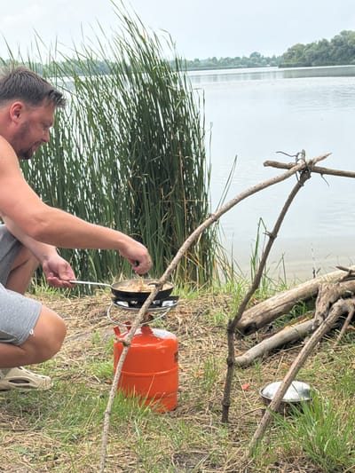 In Ruda-Siletska, Ukraine, a man is seen crouching beside a portable gas stove and orange gas cylinder, actively cooking in a pan placed on the stove. He is wearing a grey sleeveless shirt and grey shorts. The scene is set outdoors, near a body of water, with tall reeds growing along the bank. Several pieces of driftwood are visible on the ground, and a makeshift tripod made of branches stands nearby. The overall environment suggests a rustic, possibly camping, setting.