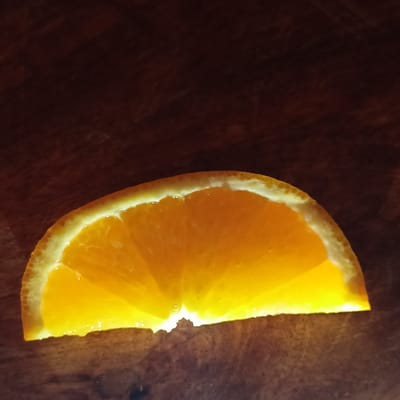 A close-up, overhead shot shows a single slice of orange fruit resting on a dark, wooden surface. The orange slice is illuminated from behind, creating a vibrant, translucent glow and casting a subtle shadow. The surface appears to be a table or countertop, with a textured, dark brown grain. The background is out of focus and a deep, shadowy brown. There are no people or actions present. The image provides no information about the city of Limerick, Ireland.