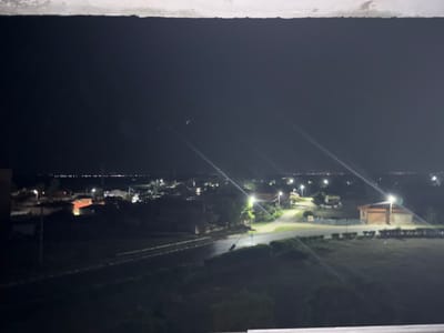 The image depicts a nighttime aerial view of a rural settlement, likely Shishmantsi, Bulgaria, characterized by a dark sky and illuminated streetlights and buildings. The foreground is dominated by a dark, possibly grassy area with a concrete barrier at the bottom edge. A road with faint lane markings is visible, winding through the scene. To the left, a few structures are partially visible, one appearing to be a multi-story building. Further into the scene, a cluster of houses with illuminated windows and roofs are scattered across the landscape. Streetlights are positioned along the roads, casting pools of light. In the distance, a faint line of lights suggests the presence of a larger town or city on the horizon. The overall impression is one of quiet, illuminated darkness over a small community at night.
