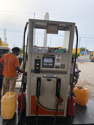 At a gas station in Thammarajupalle, India, a pump attendant wearing an orange IndianOil shirt fills a yellow jerrycan. A red fire extinguisher stands next to a fuel pump displaying a price of ₹133.33 and a volume of 69.26 liters. A second fuel nozzle is visible. In the background, a man on a motorcycle waits, and other vehicles and structures are present. The ground is paved with brick.