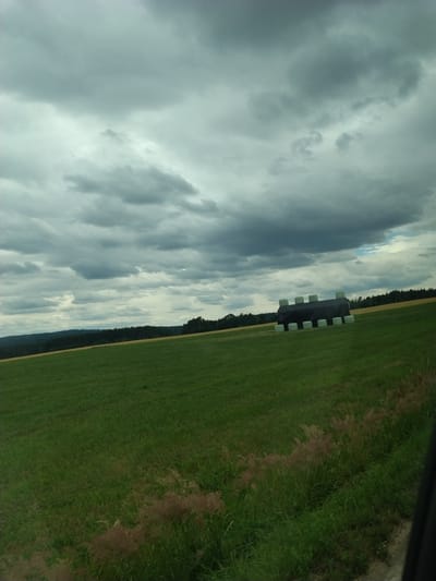 The image depicts a rural landscape under a heavily clouded sky. In the foreground, a sloped green field dominates the frame. In the middle ground, a collection of hay bales, wrapped in black plastic and some in green plastic, are arranged in a rectangular formation on the field. Beyond the hay bales, a line of dark green trees forms a dense forest that stretches across the horizon. The background consists of rolling hills and a further expanse of the forested landscape. The overall impression is one of an overcast day in a farming region. The image does not contain any people or specific identifiable city landmarks to pinpoint a location within Olešná, Czechia.