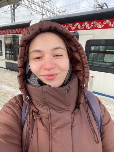 The image captures a young adult female taking a close-up selfie at a train station platform, identified as being in Fryazevo, Russia. The subject faces the camera with a slight smile, wearing a brown hooded puffer jacket and a gray and black patterned scarf or neck gaiter. A purple backpack strap is visible over her right shoulder. Behind her, a white train with a prominent red and white wavy "M" pattern, indicative of Moscow Central Diameters (MCD) rolling stock, is stationary at the platform. The platform itself is light-colored and exhibits traces of snow or ice. Overhead electrical lines and support structures are visible above the train against a cloudy sky. Bare trees are faintly discernible in the background beyond the railway.