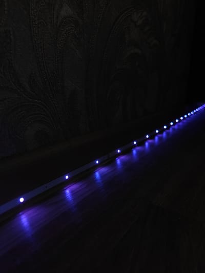 The image shows a close-up, low-angle shot of a blue LED strip light running along the base of a wall with textured wallpaper. The light strip is mounted on a white strip and extends diagonally across the frame from the bottom left corner towards the upper right. Individual blue LEDs are visible along the strip, casting a vibrant glow on the dark wooden floor. The wallpaper appears to have an ornate, swirling pattern, predominantly in dark tones, suggesting it might be nighttime or the room is dimly lit. The scene is quiet and still, with the focus on the illuminated strip as a decorative element. The overall mood is atmospheric and modern, highlighting the interplay of light and shadow in the dimly lit environment. No people or text are visible in the image. The location context is Dubovoye, Russia.