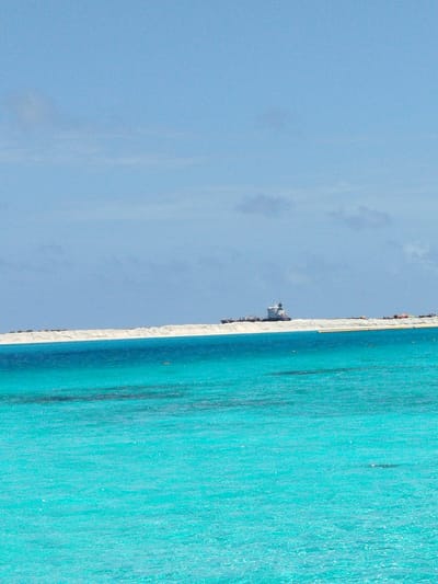 The image depicts a scenic view of a body of water with a strip of land in the background. The water is a vibrant turquoise, suggesting shallow, clear conditions, and exhibits ripples and light reflections on its surface. In the mid-ground, a distinct white embankment or artificial structure runs horizontally across the frame. On this structure, several objects are visible, including what appears to be a large vessel, possibly a ship or barge, and other smaller structures or equipment. The background consists of a clear, light blue sky with scattered wisps of clouds. The overall scene conveys a sense of coastal or marine environment. The location is identified as Gulhi Falhu, Maldives, a region known for its shallow lagoons and developing infrastructure. The image captures a moment of calm with human-made structures present in the natural landscape.