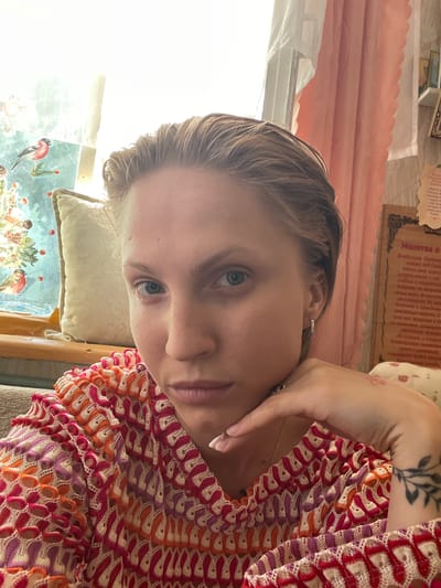 This is a close-up, rotated portrait of a young woman indoors, likely at home. She has light brown hair and is looking directly at the camera with a neutral expression. Her right arm is resting on her head, and she's wearing a colorful, crocheted top with patterns in red, pink, orange, and white. A delicate gold necklace is visible. To the left of her face, a pillow and a framed artwork with birds and greenery can be seen. Behind her, a light pink curtain hangs, and on the wall, there's a piece of paper with Cyrillic text, appearing to be a prayer. The lighting suggests it's daytime, with natural light coming from the left. The overall mood is relaxed and intimate. The visible text includes the words "Молитва о" which translates to "Prayer of."