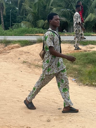 In Ganyingbo, Nigeria, three young men are walking on a dirt path parallel to a paved road. The man in the foreground, facing right, is wearing a patterned outfit with black and white stripes and green floral designs, along with black sandals. He has a shoulder bag. Behind him, a second man, also facing right, is dressed in a similar patterned outfit featuring white, green, and brown abstract designs, and is wearing dark shoes. The third man, in the background, is wearing a red and black checkered shirt and dark pants. Palm trees and dense greenery line the background, with a grey sky overhead.