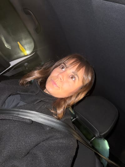 A woman with brown hair and bangs, wearing a black coat and scarf, is in the passenger seat of a car. She is wearing a seatbelt. Her eyes are wide and her mouth is slightly pursed, giving a surprised or curious expression. The interior of the car is dark, with a black ceiling and a window visible in the background. The headrest of the driver's seat is visible behind her. The lighting is somewhat dim, emphasizing the woman's face.