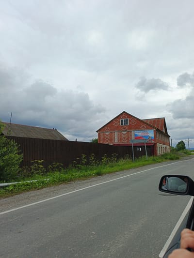 A brick building with a corrugated metal roof stands beside a road under a cloudy sky. The building has white window frames and a large banner on its side advertising "SERVICING ON CONTRACT 300,000 RUBLES 6,000,000 RUBLES" along with an image of a soldier in protective gear. A dark corrugated metal fence runs along the side of the road in front of the building, with green foliage growing behind it. The road surface is asphalt, marked with a white line. A car's side mirror reflects a person's face. The scene appears to be in Medvedyovka, Russia.