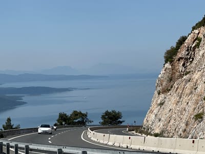 The image captures a scenic view along a winding coastal highway in Ula, Turkey. A white car is driving on the left lane, while a person on a motorbike is on the right lane of the road. The road is flanked by concrete barriers on both sides. To the right of the road, there is a steep, rocky hillside covered with sparse vegetation. To the left, there is a panoramic view of the sea with distant islands and hazy mountain ranges. The sky is clear and blue. Power lines are visible crisscrossing the landscape between the hillside and the sea.
