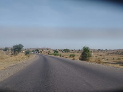The image shows a paved road stretching out into the distance, with a few scattered trees and shrubs on either side. The road is empty with no cars or people visible. The environment is a rural area, with a dry, brown landscape under a clear, bright blue sky. The road appears to be straight and leads into a valley, with low hills in the distance. The location is in Ture, Nigeria, at the coordinates latitude: 9.83, longitude: 11.375.