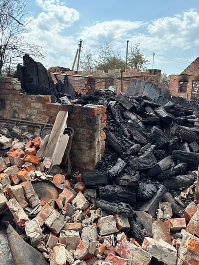 This image depicts the extensive remains of a structure, indicative of severe destruction by fire, located in Nedryhailiv, Ukraine. The scene is devoid of human presence or activity.

In the foreground, a large pile of fragmented, reddish-brown and gray bricks is visible, interspersed with smaller pieces of debris and some bent metallic elements. Adjacent to this, centrally positioned, stands a partially collapsed brick wall, approximately a meter high, with visible scorch marks. Leaning against this wall is a roughly rectangular, light-colored piece of material, possibly concrete or plasterboard.

To the right of the partial brick wall, and extending into the midground, is a substantial accumulation of uniformly blackened, heavily charred wooden timbers and smaller debris fragments, suggesting burnt structural elements and furnishings.

In the background, additional remnants of brick walls and structures are present, also exhibiting damage. Bare deciduous trees rise behind these ruins, and two utility poles with overhead power lines are visible against a partly cloudy blue sky.
