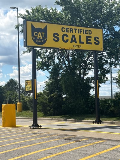 The image shows a sign for "CAT SCALE CERTIFIED SCALES" in Burbank, California. The sign is yellow with black lettering and a black cat logo. It is mounted on two black posts. There is a smaller sign on one of the posts that also says "CERTIFIED SCALE". The sign is located in a parking lot with yellow lines painted on the asphalt. There are trees and a street light in the background. The sky is blue with some clouds. There are no people or animals visible in the image.