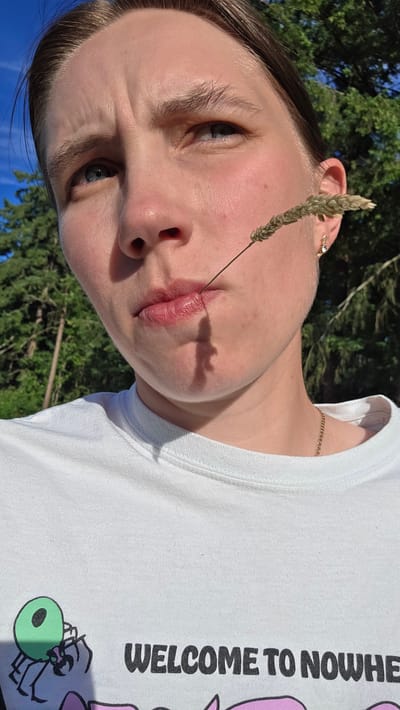 A close-up, upward-angled shot captures a person's face and upper torso. The person has light skin, brown hair pulled back, and blue eyes. A stalk of dried grass is positioned horizontally across their lips, with the tip resting on their lower lip. Their expression is neutral, with a slight furrow in their brow. They are wearing a white t-shirt with a graphic design on the front, featuring a green cartoon spider and the words "WELCOME TO NOWHERE" printed in black lettering. A thin gold chain necklace is visible around their neck. The background consists of a blurry mix of green trees and a bright blue sky, suggesting an outdoor setting, likely in a park or wooded area. The lighting indicates it is daytime, possibly late afternoon due to the shadows. There is no indication of the specific location within Portland, United States.