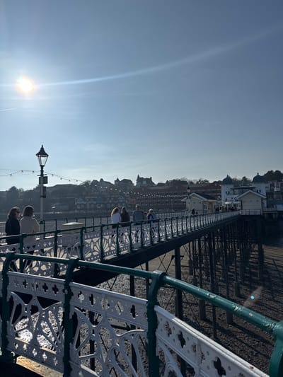 The image captures Penarth Pier in Penarth, United Kingdom, on a clear, sunny day. The sun is positioned in the upper left, producing lens flare and a horizontal light streak across the blue sky; a faint contrail is visible.
The pier's structure dominates the lower and mid-sections of the frame, featuring dark green structural elements and white, ornate cast-iron railings with a dark green top rail. The ground beneath the pier is a dry, rocky beach or seabed, indicating low tide, with visible shadows cast by the pier's support piles.
Approximately nine individuals are present on the pier. In the foreground-left, two women walk away from the camera, one in a dark top, the other in a light beige top. Further along the pier, a group of five people are walking or standing, mostly facing away. Two additional individuals are visible further down the pier. A black, ornate street lamp post stands on the left side of the pier. A string of decorative lights is strung below the lamp post and extends further along the pier.
In the mid-ground, along the pier's length, several small white buildings with pitched roofs are visible, leading to a larger white building with a distinctive domed roof at the pier's far end. The background displays a coastal town built on a hillside, featuring numerous multi-story buildings with varied architectural styles and rooflines, some with darker facades. Additional string lights are visible stretching along the waterfront in the background, connecting to the town structures.