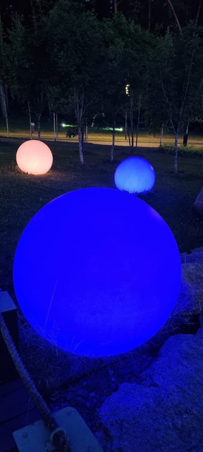 At night in Shulgino, Russia, three spherical illuminated objects are displayed on a grassy area with scattered trees and rocks. The largest sphere in the foreground emits a vibrant blue light. A second sphere, smaller and to the left, glows with a soft pinkish-orange hue. A third sphere, also smaller, is positioned behind the pink one and emits a light blue glow. The area is dark, with the illuminated spheres providing the primary light source. A portion of a wooden structure and a thick rope are visible in the lower left corner of the frame. The background shows a line of trees and what appears to be a pathway or road with distant lights.