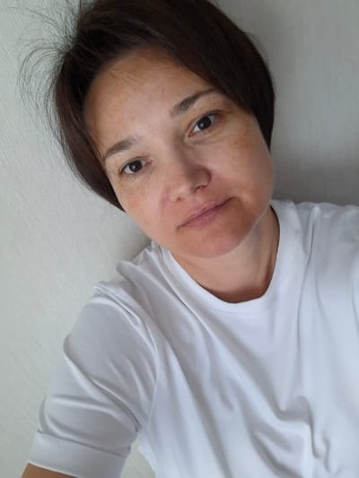 This is a close-up, selfie-style photograph of a person lying down. The person has short, dark brown hair and appears to be wearing a plain white t-shirt. The background is a light-colored, textured wall.

The subject's face is partially visible, angled towards the camera. They have fair skin with visible freckles and some redness on their cheeks. Their eyes are dark brown and they appear to be looking directly at the camera. The lighting suggests it is daytime, possibly indoors. There are no explicit indicators of the time of day or weather conditions beyond the natural light. The setting appears to be a simple, indoor environment. The subject's expression is neutral, with a slight hint of a smile. There is no visible text in the image. The location context is Svobodny, Russia.