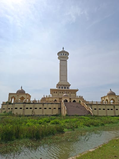 This landscape-oriented image captures a grand, ornate building, likely a mosque or a structure influenced by Islamic architecture, situated beside a body of water. The central feature is a tall, slender, minaret-like tower with a domed top, constructed from a light-colored material, possibly beige or cream stone.

The main building extends horizontally across the frame, featuring multiple smaller domes or cupolas along its roofline and upper levels. It displays intricate architectural details, including arched openings, balconies with decorative railings, and multi-storied sections. The structure appears substantial and well-crafted.

Between the building and the body of water—which appears to be a river or canal—is a wide strip of lush, dense green vegetation, including bushes and grass. The water's surface shows gentle ripples, reflecting the bright, pale blue sky, which has a few faint, wispy clouds. The lighting suggests it is daytime, with a soft illumination, indicating either morning or late afternoon. There are no people visible, contributing to a serene and peaceful atmosphere. The architectural style aligns with the provided location context of Kruengmate, Indonesia.