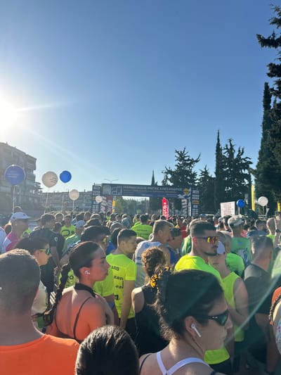 A photograph captures a large crowd gathered in the Stavroupoli Municipal Unit, Greece, for the "Marathon Megas Alexandros" race. The participants are standing in a dense group, many wearing brightly colored green and orange shirts. The crowd appears to be lining up at the starting line of the race, marked by a banner overhead. The banner indicates the event is a health run and dynamic walking event of 10,000 meters, scheduled for April 28, 2020. Various sponsors and logos are displayed on the banner, including "bwin," "MIN," and "WH."

The scene is filled with balloons, some of which are branded with the name "ΔΗΜΟΣ ΑΜΠΕΛΟΚΗΠΩΝ." Trees and buildings form the backdrop of the event. The sun is high in the sky, casting strong light across the scene.