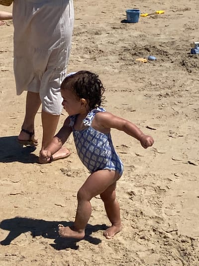 The image shows a young child in a blue and white patterned swimsuit on a sandy beach, likely in Conil de la Frontera, Spain. The child is in a dynamic pose, leaning forward with one arm raised and clenched into a fist, appearing to be mid-movement or play. Their legs are bent, with one foot visible in the sand. Another person, partially visible from the waist down, is wearing a white garment and sandals, and is reaching out towards the child, possibly helping them or playing with them.
The setting is a beach with the sand showing textures and indentations, suggesting it might be wet or recently disturbed. There are some small beach toys scattered on the sand in the background, including a blue bucket and a yellow shovel. The sunlight casts a strong shadow of the child and the other person on the sand, indicating it is likely a sunny day with the sun relatively high in the sky, suggesting midday. The overall mood appears to be one of active, playful engagement, and possibly a parent or guardian interacting with a child on a beach outing. No visible text is present in the image.