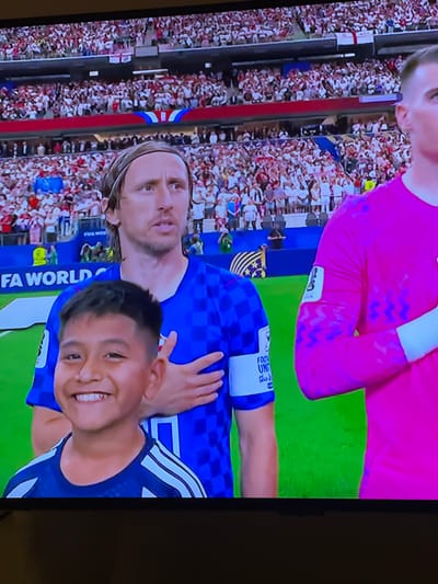 The image shows a close-up of a soccer match with a large crowd in the background. In the foreground, a young boy with dark hair and a wide smile stands in front of two soccer players. The boy is wearing a dark blue jersey with white stripes on the shoulders. The player on the left is wearing a blue and white checkered jersey, and the player on the right is wearing a bright pink jersey. Both players have their hands on their chests, presumably during a national anthem. The background shows a stadium filled with spectators wearing white and red attire, with some flags visible. The overall setting is a soccer field with a large audience. The image does not provide any information about the city of Florești, Romania.