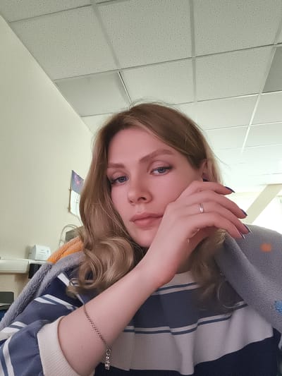 A young woman with blonde wavy hair is sitting indoors. She is wearing a striped sweater and has a blanket draped over her shoulders. Her left hand is raised to her face, and she has a ring on her ring finger. Her right arm is visible and has a bracelet. The room appears to be an office or a similar institutional setting, with a ceiling with square panels and a wall in the background. The woman's gaze is directed towards the camera. The city of Postol, Russia is not identifiable as a feature of this image.