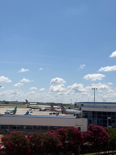 The image captures an airport scene with several airplanes parked at gates outside a modern terminal building. The terminal features a glass facade reflecting the sky and clouds. Tall light poles with multiple lamps are visible along the tarmac. Lush green trees with vibrant pink flowers frame the right side of the image. The sky is a clear blue with scattered fluffy white clouds, suggesting a pleasant day. The overall impression is one of activity and routine at a busy airport.