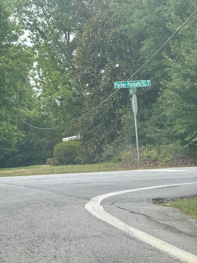 A daytime street scene shows a road sign for "Parker-Pucketts WAY SW 5000," which indicates the location of Parker-Pucketts Way Southwest, number 5000. The sign is green with white lettering and includes a directional arrow. Adjacent to the sign is dense green foliage from trees and undergrowth. The road is gray asphalt with a white lane marker. The location is in Mountain Park, United States of America.