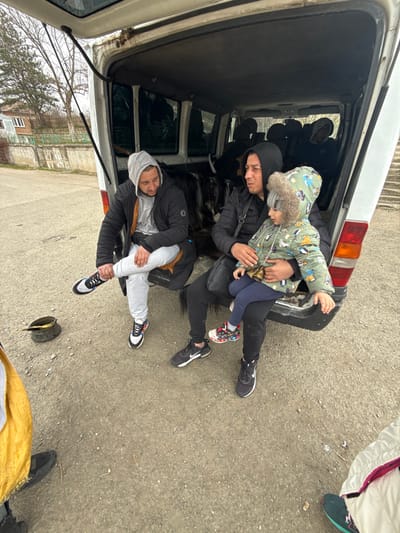 A white panel van with an open rear hatch is parked on an unpaved surface in Zidarovo, Bulgaria. Two adult males and a small child are seated on the edge of the van's rear threshold. The male on the left wears a grey hooded sweatshirt, a black puffer jacket, light grey sweatpants, and black and white sneakers, with his right leg crossed. The male on the right wears a dark hooded top, a black puffer jacket, dark pants, and black sneakers, with a dark shoulder bag. He holds a young child dressed in a multi-colored hooded winter jacket, dark blue pants, and colorful sneakers on his lap. Additional obscured figures are visible within the darkened interior of the van. On the ground to the left, a dark metal bucket or container is present. The background features a light-colored house with a fence and bare trees under an overcast sky. Partial views of two other individuals' clothing and footwear are visible in the immediate foreground, outside the van.