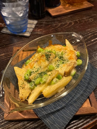A glass dish containing a baked potato dish with edamame and herbs is sitting on a blue and white striped fabric napkin, which is on a wooden tray. The dish is surrounded by a wooden surface. There is a glass of ice water in the lower left corner of the image. The coordinates 34.458, 133.995 correspond to a location in Naoshima, Japan, which is known for its art installations and museums. This image does not depict an art installation or museum.
