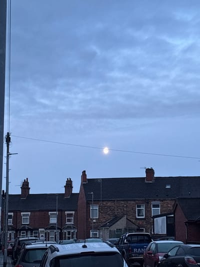 The image depicts a quiet residential street scene under an overcast sky, likely taken during early morning or late afternoon given the diffused light. The sky, occupying the left side of the frame, is a uniform light blue-grey with subtle cloud formations. A bright, circular orb, presumably the sun or moon obscured by clouds, is prominent in the upper-right portion of the sky.
On the right side of the image, a row of terraced or semi-detached houses is visible. These buildings are constructed from dark red brick with dark, possibly slate or tile, roofs. Multiple traditional brick chimneys punctuate the rooftops, and several windows with white frames are visible on the house facades.
Along the street in front of the houses, several vehicles are parked. A large white van is prominent in the foreground on the far right. Behind it, a blue van displays the partial text "RAN" on its side, and a dark-colored car is also visible. In the bottom-left foreground, a utility pole with multiple wires extends upwards, while a single, thin wire runs diagonally across the sky from the lower-left, passing near the bright orb.
The scene conveys a calm, subdued atmosphere, typical of a cloudy day in a residential area. The architecture and street layout are consistent with the provided location context of Stoke-on-Trent, United Kingdom.