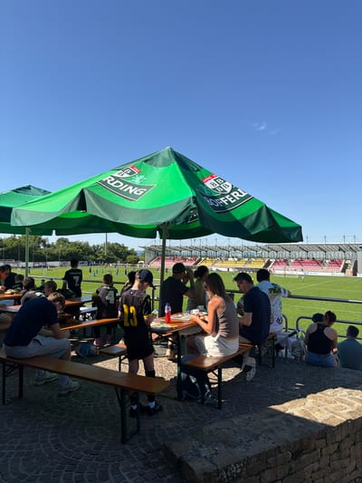 The image shows a group of people sitting at tables under green umbrellas. There are two rows of tables, with people sitting on benches facing the soccer field. On the field, a soccer game is taking place. In the background, a stadium with red and yellow seats is visible. This scene is likely in Fentange, Luxembourg, as indicated in the prompt.