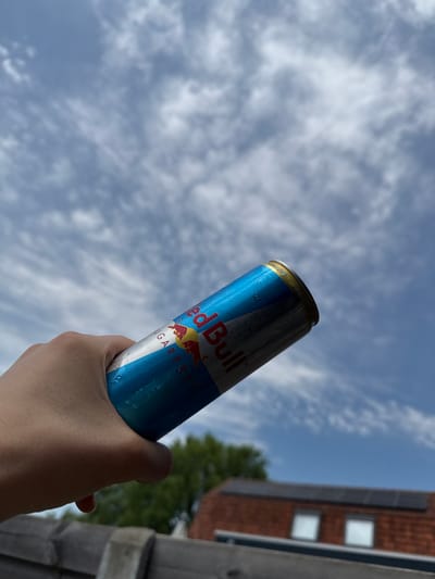 A hand holds a Red Bull can against a cloudy sky. The can is blue with a silver bottom and has water droplets on its surface. The Red Bull logo, featuring two red bulls, is visible. Text on the can indicates "SUGAR FREE". In the background, the roof of a house with solar panels and some greenery are discernible. The scene appears to be outdoors, possibly in a residential area.